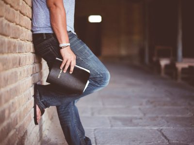 man holding Holy Bible leaning on bricked wall